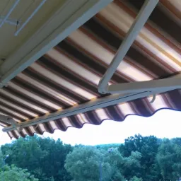 Zewnętrzna markiza w brązowo-białe pasy zamontowana nad balkonem, widok na drzewa w tle.