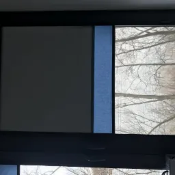 Okno z roletą dzień noc: część zasłonięta szarą tkaniną, część z niebieskim pasem, widok na drzewa bez liści za oknem.