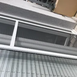 Zamknięta moskitiera plisowana w białej ramie, widok z dołu na tle szarego tynku.