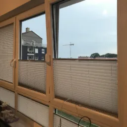 Okna z zamontowanymi plisowanymi osłonami okiennymi w kolorze kremowym, widok na budynek mieszkalny i żuraw budowlany w tle.