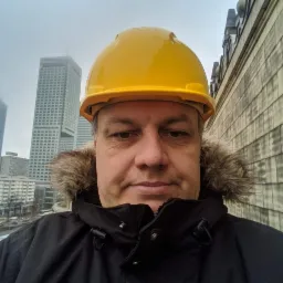 Portret instalatora w żółtym kasku na tle miejskiego krajobrazu z wysokimi budynkami w mglisty dzień. Ujęcie z dołu, widoczna część budynku.