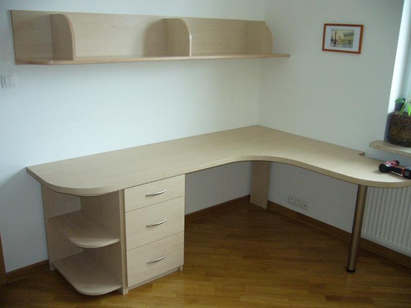 Jasny, narożny biurko z trzema szufladami i półkami, uzupełnione wiszącą półką nad nim, w pokoju z parkietem i grzejnikiem przy oknie.