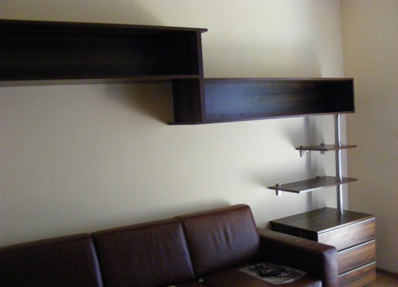 Ciemnobrązowe, asymetryczne półki wiszące na ścianie w Łomiankach, nad brązową skórzaną sofą. Obok widoczny regał z trzema półkami na metalowym stelażu.