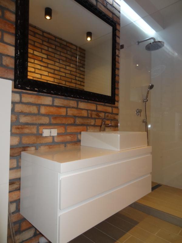 Nowoczesna łazienka z białą szafką pod umywalkę z szufladami i prostokątną umywalką nablatową, na tle ściany z czerwonej cegły i lustra w ozdobnej czarnej ramie, obok kabiny prysznicowej.