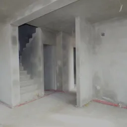 Wnętrze w trakcie prac tynkarskich: surowe ściany i schody pokryte świeżym tynkiem, widoczne kable instalacji elektrycznej przy podłodze.