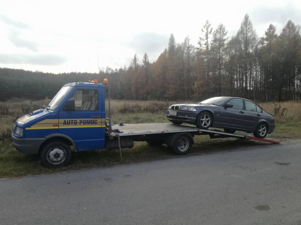 Niebieska laweta z napisem 'AUTO POMOC' transportuje szary samochód osobowy na tle jesiennego krajobrazu z drzewami.