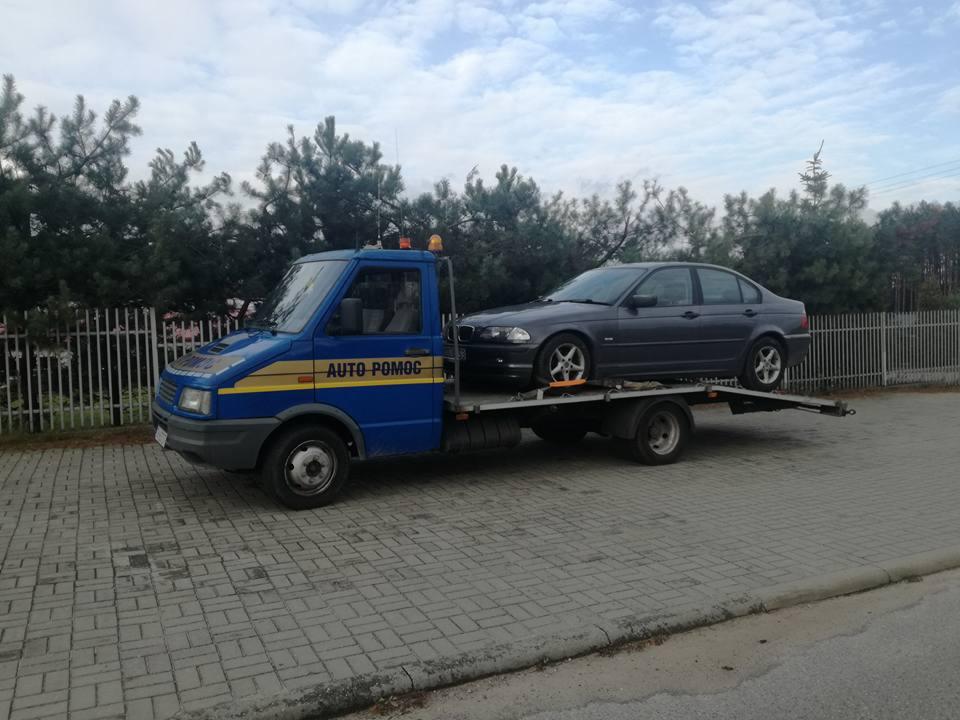 Niebieska laweta z napisem 'AUTO POMOC' przewozi szary samochód osobowy na tle drzew i szarej kostki brukowej.