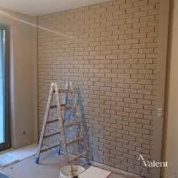 Ciszyński - Ściana wykończona tapetą imitującą cegłę w odcieniach beżu, z widoczną drewnianą drabiną i wiadrem z farbą, sugerującą świeżo zakończone prace malarskie.