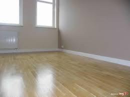 Pusty pokój z parkietem w odcieniach brązu, świeżo pomalowane ściany w kolorze beżowym, białe listwy przypodłogowe i grzejnik pod oknem. Widoczne refleksy światła na podłodze.