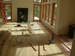 Surowe drewno podłogowe rozłożone na podłodze w jasnym, przestronnym pokoju z dużymi oknami, przygotowane do montażu, widoczne narzędzia i materiały budowlane.