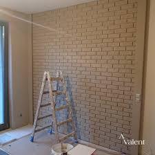 Ściana wykończona tapetą imitującą cegłę w odcieniach beżu, z widoczną drewnianą drabiną i wiadrem z farbą, sugerującą świeżo zakończone prace malarskie.