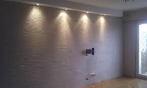 Ściana w pokoju z dekoracyjną fakturą i oświetleniem punktowym, widoczny uchwyt na telewizor i gniazdko elektryczne.