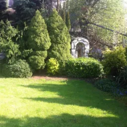 Zadbany ogród z równo przystrzyżonym trawnikiem, ozdobnymi krzewami, iglakami i kamienną grotą z rzeźbą w tle.