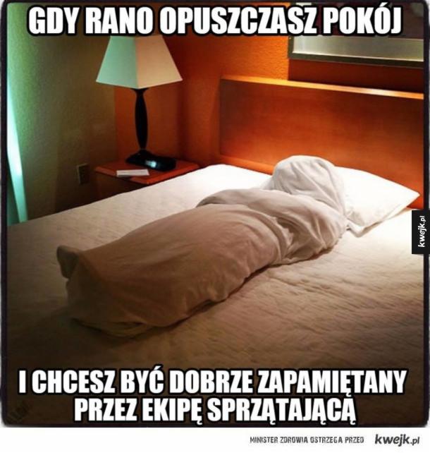 Łóżko w pokoju hotelowym z kołdrą uformowaną w kształt ciała, przygotowane dla ekipy sprzątającej, z lampką nocną na szafce.