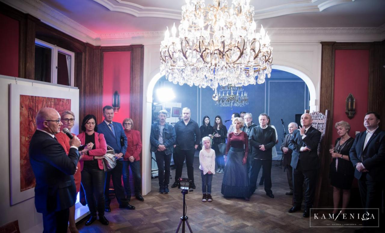 Wystawa obrazów w galerii: mężczyzna w garniturze przemawia do zgromadzonych gości na tle abstrakcyjnego obrazu i kryształowego żyrandola.