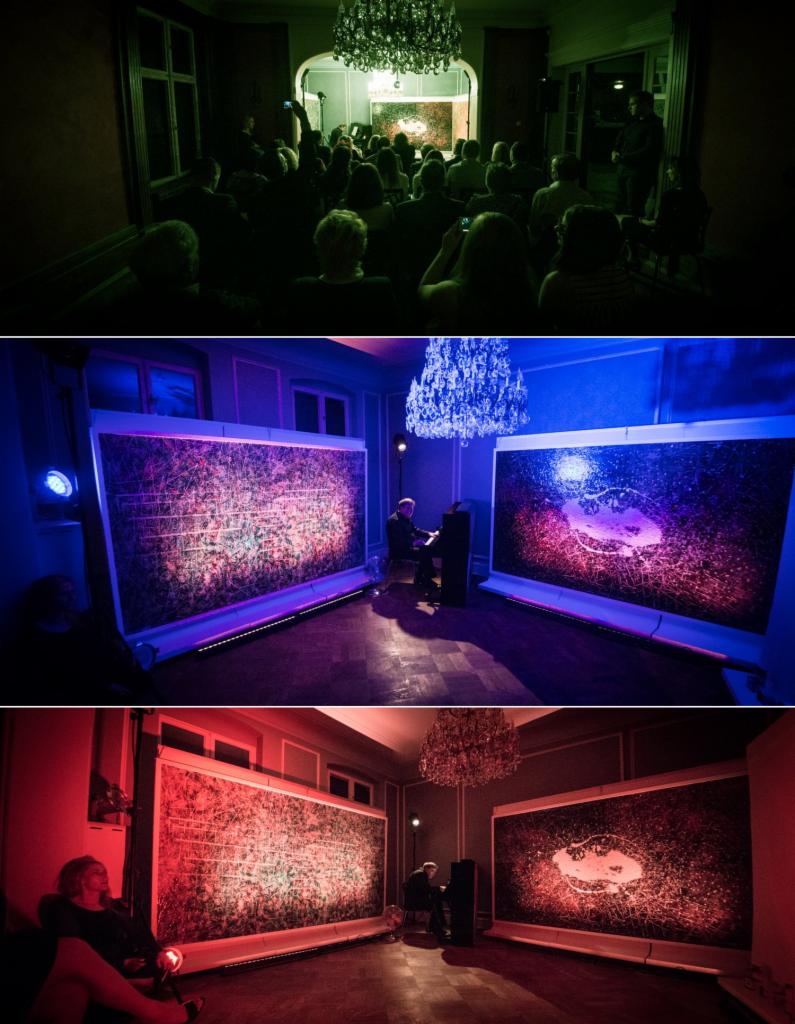 Wystawa malarstwa artystycznego w pomieszczeniu z kryształowym żyrandolem, oświetlona kolorowymi światłami, z pianinem i widownią.