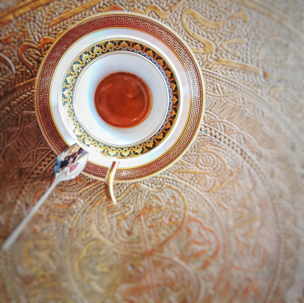 Filiżanka espresso na bogato zdobionej podstawce i tle z wzorem w stylu Versace, z odłożoną srebrną łyżeczką po lewej stronie.