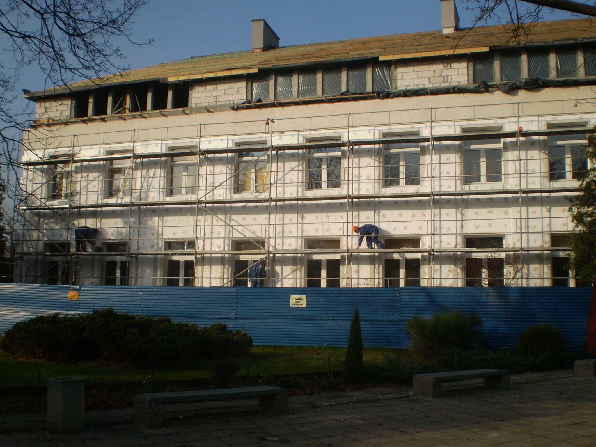 Budynek wielopiętrowy w trakcie prac elewacyjnych: rusztowania, pracownicy w kombinezonach, częściowo ocieplona fasada, zabezpieczenia na dachu, widoczny teren budowy z niebieskim ogrodzeniem.