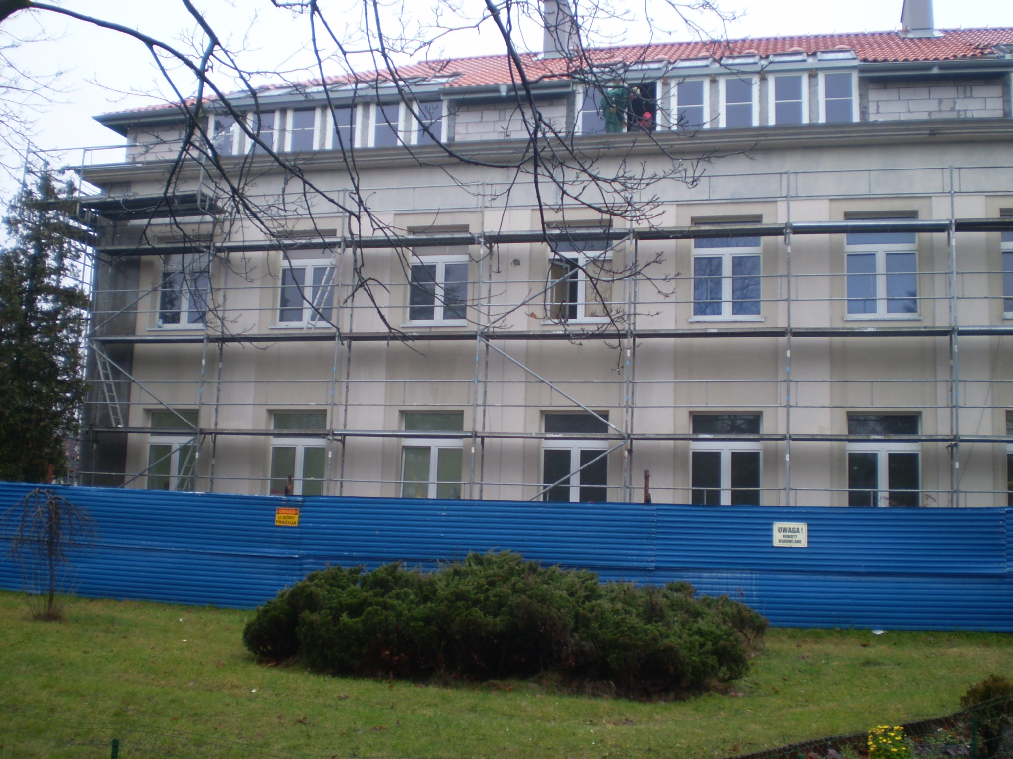 Budynek w trakcie prac elewacyjnych, widoczne rusztowania, niebieskie ogrodzenie i pracownicy przy oknach na poddaszu.