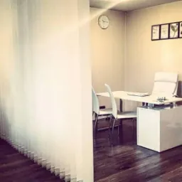 Gabinet dietetyczny z urządzeniem LPG, biurkiem z krzesłami, zegarem na ścianie i certyfikatami w ramkach.