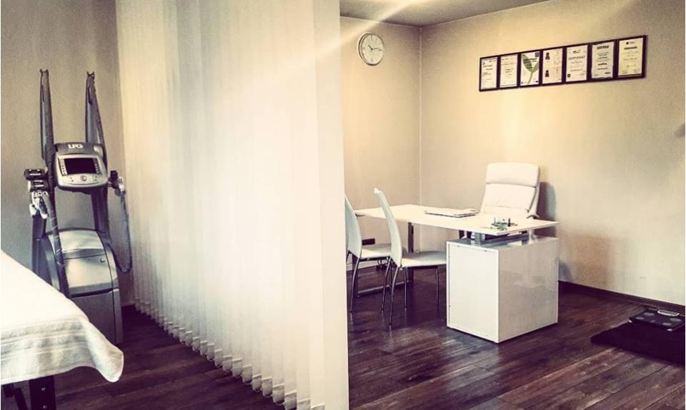 Gabinet dietetyczny z urządzeniem LPG, biurkiem z krzesłami, zegarem na ścianie i certyfikatami w ramkach.