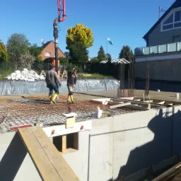 Budowa piwnicy z gotowych elementów: betonowe ściany szalunkowe, stalowe zbrojenie, dwóch robotników w żółtych gumowcach na tle błękitnego nieba i pompy do betonu.