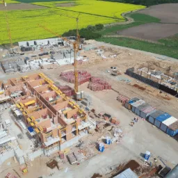 Widok z góry na plac budowy z kilkoma budynkami w różnym stadium realizacji, z widocznymi fundamentami, murowanymi ścianami z czerwonej cegły, szalunkami, żurawiami budowlanymi i kontenerami...