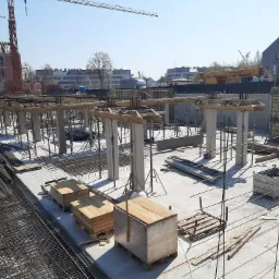 Widok z g&oacute;ry na plac budowy z przygotowanymi fundamentami pod budynek, widoczne zbrojenia, betonowe słupy, drewniane szalunki i dźwigi w tle.