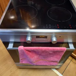 Czarna płyta indukcyjna i piekarnik z wyświetlaczem pokazującym godzinę 14:24, z różowym ręcznikiem wiszącym na uchwycie piekarnika.