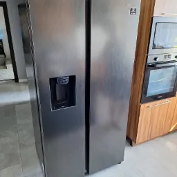 Dwudrzwiowa lodówka Samsung z dystrybutorem wody i kostkarką, widoczna obok zabudowanej szafki z piekarnikiem, na tle szarej podłogi.