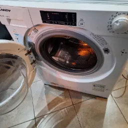 Biała pralka marki Candy z otwartymi drzwiczkami, widoczny bęben z metalowymi otworami, panel sterowania z wyświetlaczem i pokrętłem, plamy i zabrudzenia na obudowie, zdjęcie z bliska na tle płytek...