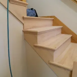 Jasne, drewniane schody w trakcie montażu, widoczne niedokończone stopnie i fragment poręczy, niebieski kabel wzdłuż ściany.