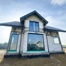 Dom w trakcie budowy z zamontowanymi oknami o czarnych ramach, widoczny z perspektywy żabiej, na pierwszym planie piasek, w tle osoba w czerwonej czapce.