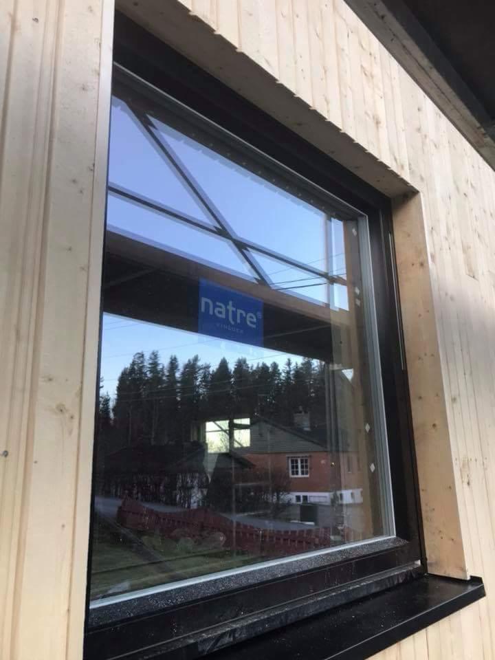 Zamontowane okno z czarną ramą w drewnianej elewacji, na szybie widoczna naklejka producenta i odbicie krajobrazu z domem.