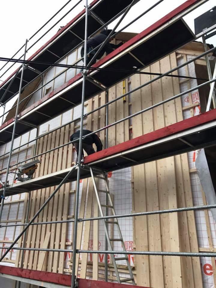 Montaż drewnianej elewacji zewnętrznej domu z rusztowania, widoczne deski, izolacja i pracownicy.