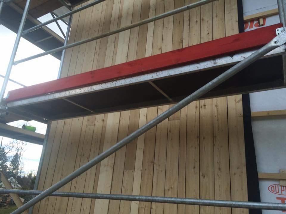 Montaż drewnianej boazerii na elewacji budynku, widoczne rusztowanie i folia izolacyjna.