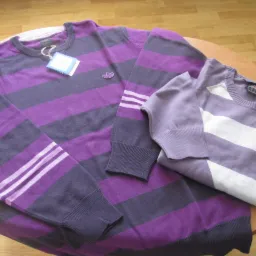Dwa swetry w paski, jeden fioletowo-granatowy z logo Adidas na piersi i trzema paskami na ramieniu, drugi liliowo-biały, oba z metkami, leżące na okrągłym stole.