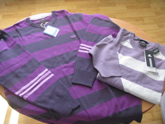 Dwa swetry w paski, jeden fioletowo-granatowy z logo Adidas na piersi i trzema paskami na ramieniu, drugi liliowo-biały, oba z metkami, leżące na okrągłym stole.