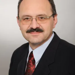 Portret mężczyzny w okularach, z wąsami, w ciemnym garniturze i czerwonym krawacie na szarym tle.