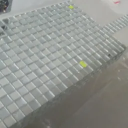 Ułożona mozaika z małych, kwadratowych płytek w odcieniach bieli i szarości, z żółtymi naklejkami na niektórych płytkach, widoczna puszka elektryczna w ścianie i fragment folii ochronnej.