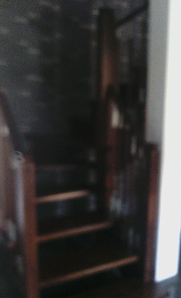 Nierówne drewniane schody w słabo oświetlonym wnętrzu, widoczne fragmenty ciemnej tapety w tle.