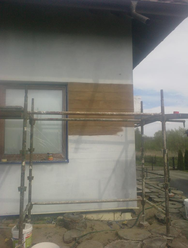 Elewacja domu w trakcie malowania na biało, fragment pomalowany na brązowo, widoczne rusztowanie i zabezpieczone okno.