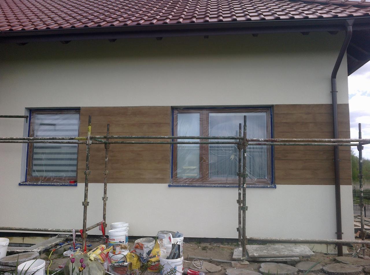 Elewacja domu jednorodzinnego w trakcie wykańczania, widoczne rusztowania, okna zabezpieczone folią, dolna część ściany pomalowana na biało, górna imitująca drewno, puszki farb i narzędzia na ziemi.