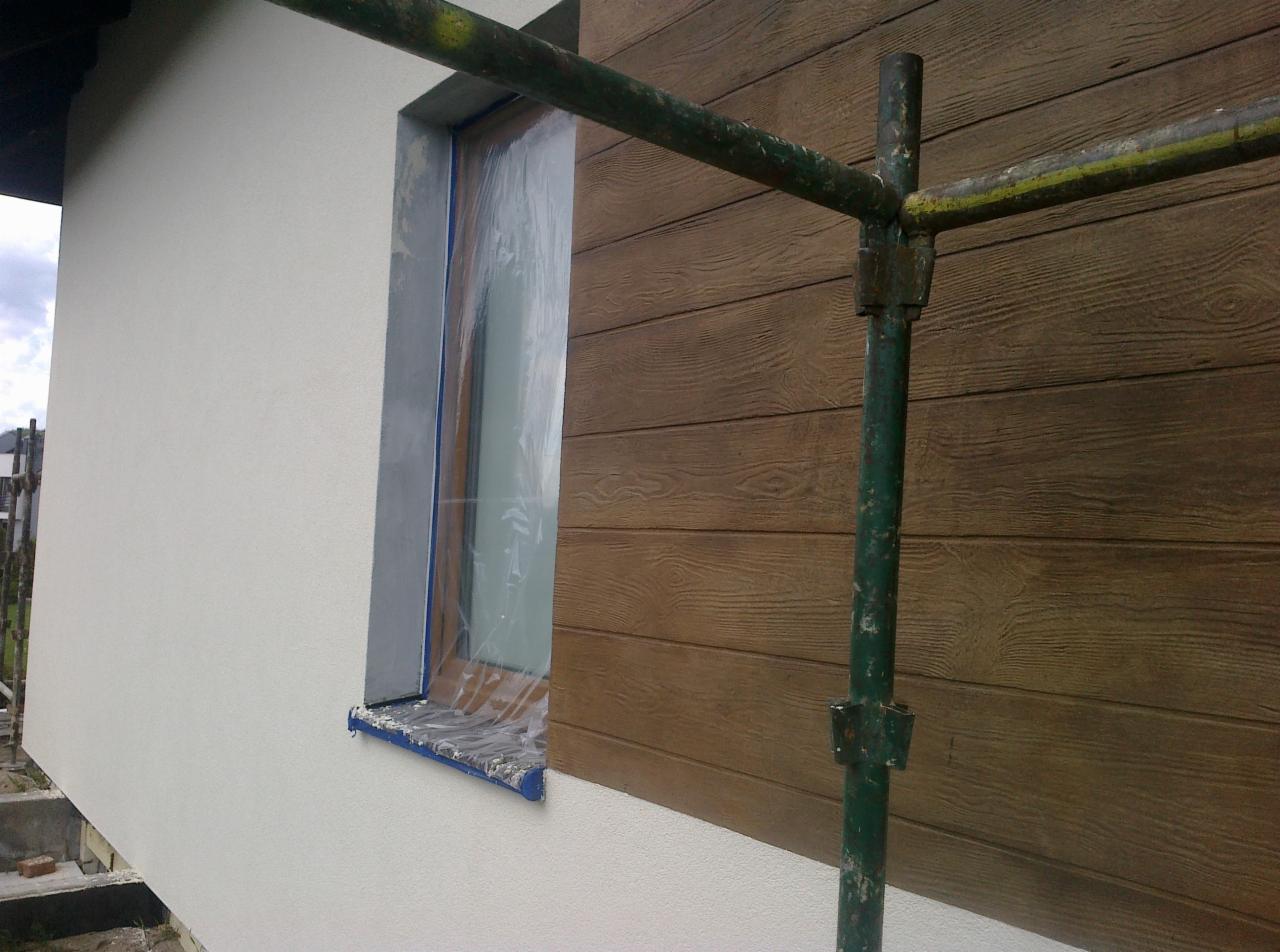 Fragment elewacji budynku z oknem zabezpieczonym folią malarską, obok fragment rusztowania i ściana wykończona imitacją drewnianych desek.
