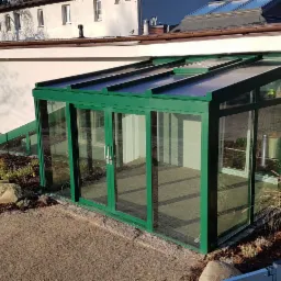 Zewnętrzna zabudowa tarasu z zielonymi profilami aluminiowymi i przesuwnymi drzwiami, widok z zewnątrz na trawnik i budynek z oknami dachowymi.