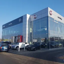 Duży, nowoczesny salon samochodowy z aluminiowymi oknami i logo marek Fiat, Alfa Romeo i Jeep, prezentujący samochody na zewnątrz i wewnątrz budynku.