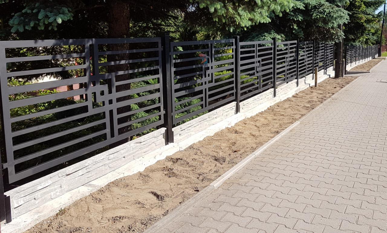 Nowoczesne, spawane ogrodzenie z metalowymi przęsłami o geometrycznym wzorze, osadzone na podmurówce z imitacji kamienia, widoczne z perspektywy chodnika.