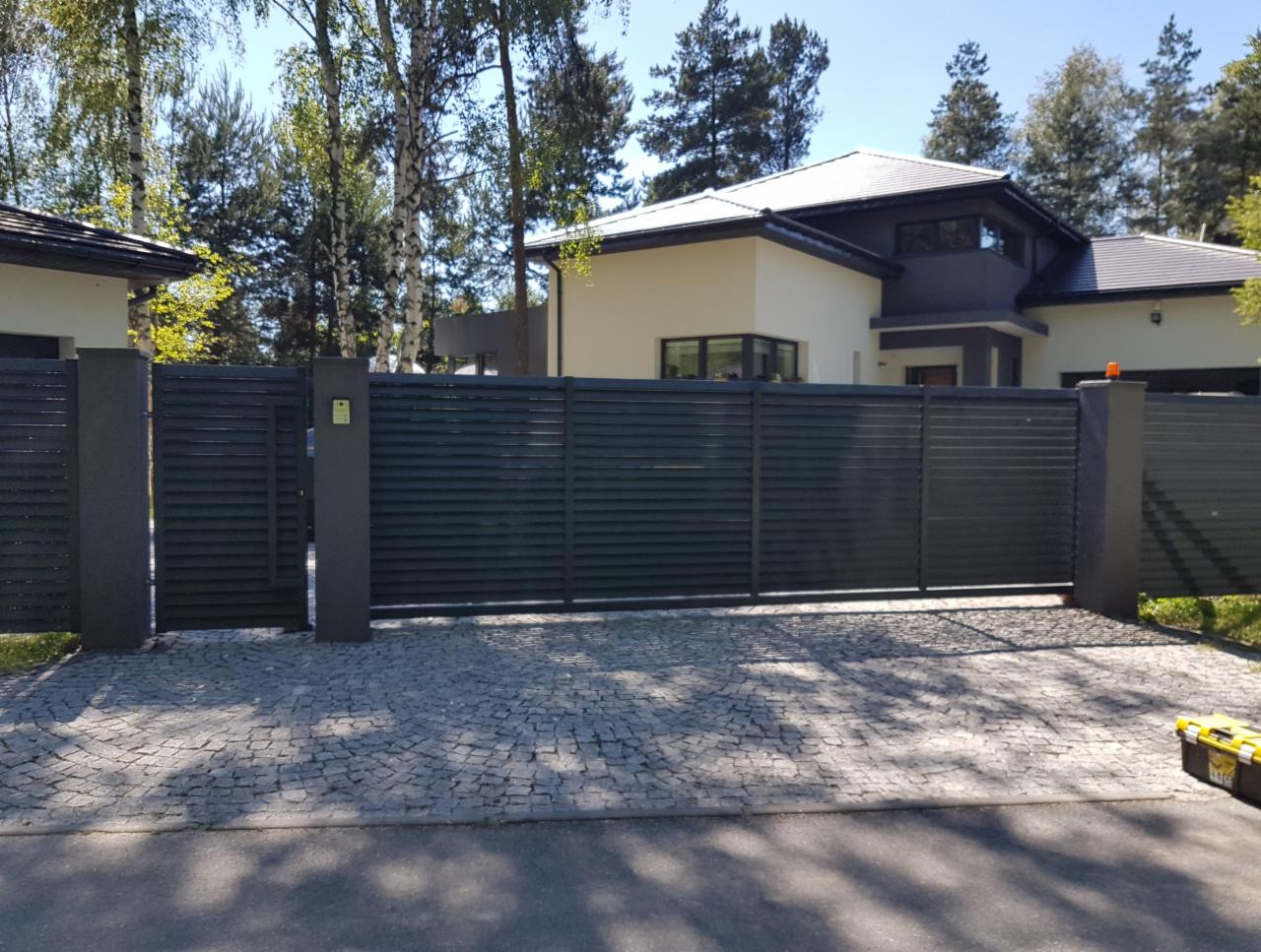 Nowoczesna, pozioma brama przesuwna z szarych, metalowych profili na tle domu z jasną elewacją i brukowanym podjazdem z kostki granitowej.