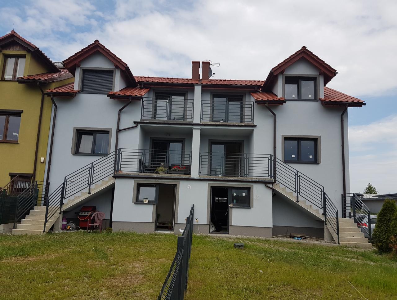 Dwupiętrowy budynek z balkonami i zewnętrznymi schodami z czarnymi metalowymi balustradami, widok z poziomu trawnika.