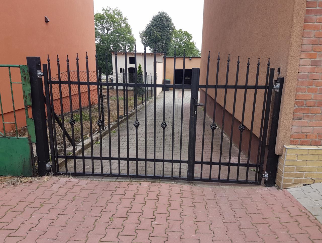 Czarna, metalowa brama z dekoracyjnymi elementami, zamontowana między dwoma budynkami, widok z zewnątrz na brukowany podjazd.